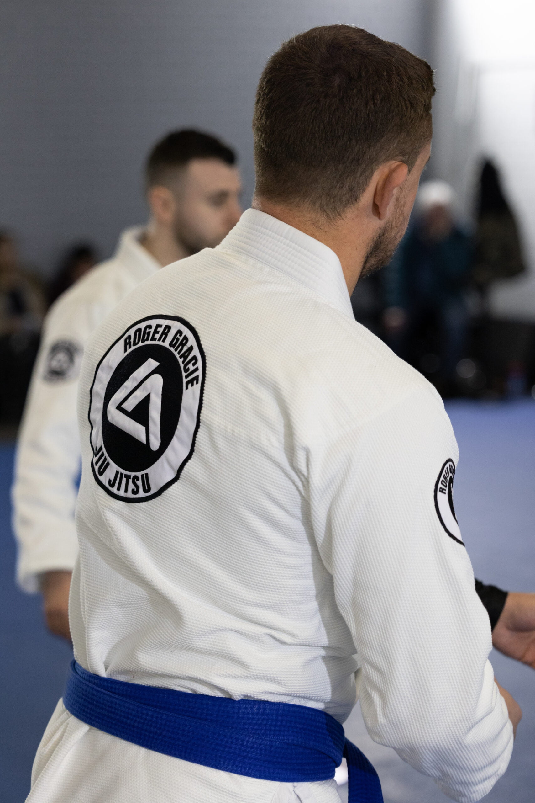 The back of a Roger Gracie blue belt assistant coach overseeing the kids train.